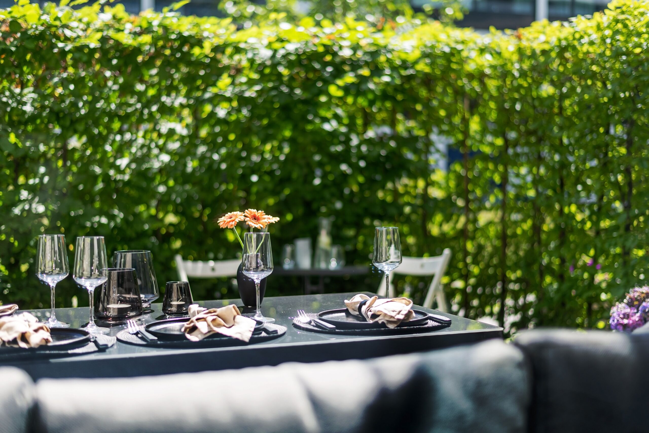 Outdoor dining table set with wine glasses, napkins and a floral centerpiece against a lush garden backdrop